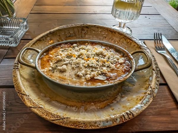Obraz Delicious Hot Casserole with Feta Cheese Served in a Rustic Pan at an Outdoor Restaurant. Al Fresco Dining: A Hearty Baked Dish on a Wooden Table at a Summer Cafe Terrace