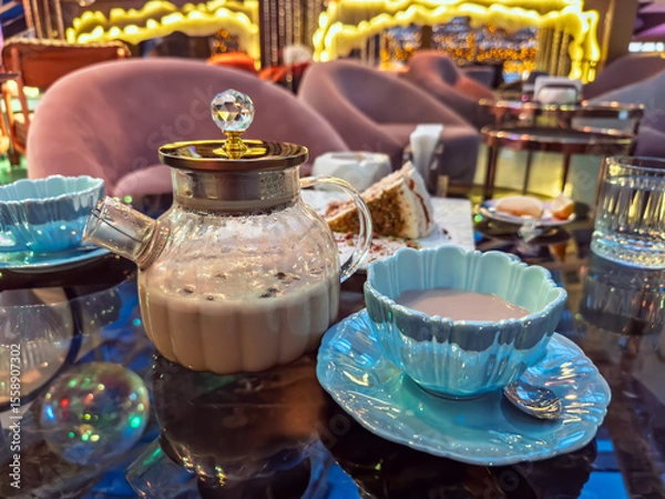 Obraz A chic tea time setup with a glass teapot, elegant blue cup, and dessert in elegant cafe.Elegant glass teapot and a stylish blue cup of milk masala tea served in a modern, upscale restaurant lounge