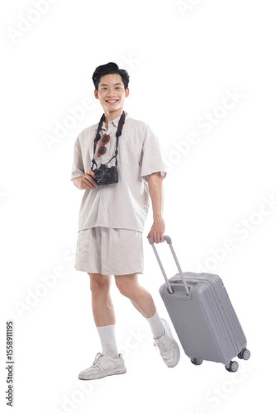 Fototapeta Ready for Adventure: A smiling young man, ready for his journey, pulls a grey suitcase, camera and sunglasses hanging around his neck.