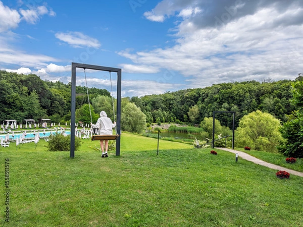 Obraz Girl in a hoodie relaxing on a large modern swing at a summer resort overlooking a pool and green forest.Summer leisure and recreation concept. A person rests on a swing in a beautiful resort area
