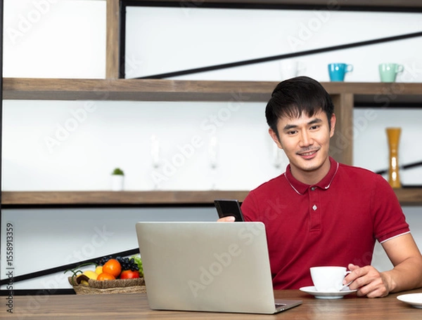 Fototapeta Young Smiling handsome wearing red t-shirt meeting online and drinking a cup of coffee and
work from home. Communicating online, writing emails, distantly working or studying on computer.