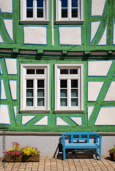 Fototapeta Facade of half timbered house and blue bench in Idstein Germany. Green and white wall and truss with windows of a medieval old house. Idstein is a idyllic small town near Frankfurt.