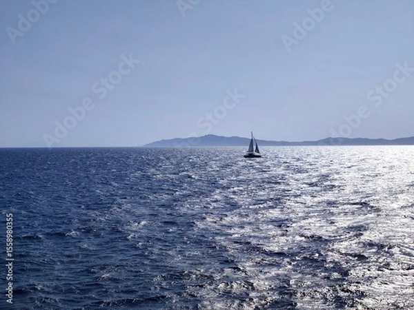 Fototapeta Photo of the Aegean Sea under a brilliant summer sky, golden sunlight dancing on the waves, while in the distance a solitary sailboat drifts peacefully across the shimmering blue horizon.