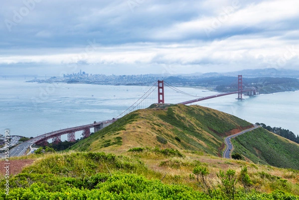 Fototapeta Panoramic view of the Golden Gate Bridge spanning San Francisco Bay with cityscape, lush hills, and boats, captured from a high vantage point on a cloudy spring day