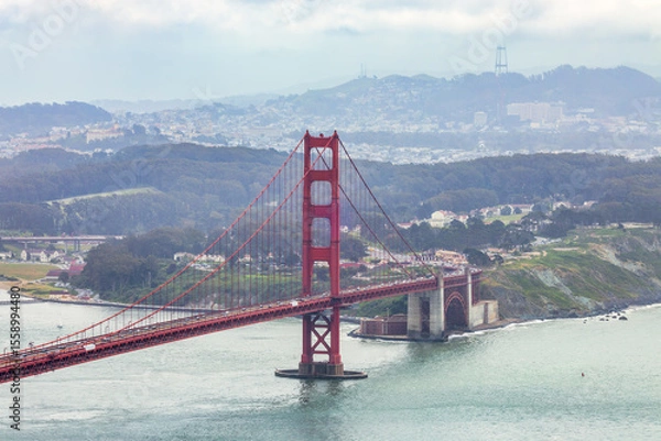 Fototapeta Panoramic view of the Golden Gate Bridge spanning San Francisco Bay with cityscape, lush hills, and boats, captured from a high vantage point on a cloudy spring day