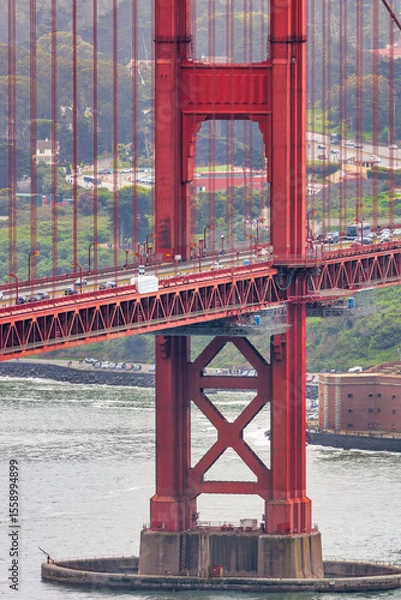 Fototapeta Close-up of Golden Gate Bridge tower with Palace of Fine Arts in the background, San Francisco cityscape and boats on the bay visible through the iconic red structure.