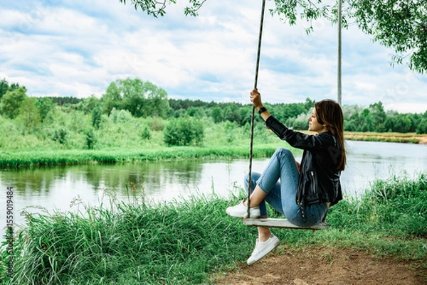 Fototapeta A happy young girl is sitting on a swing in a beautiful tree by the river.