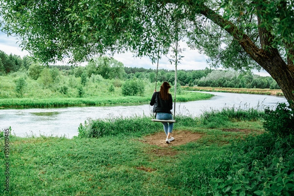 Fototapeta A happy young girl is sitting on a swing in a beautiful tree by the river.
