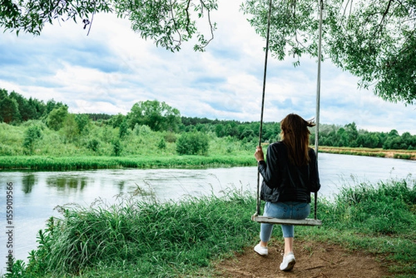 Fototapeta A happy young girl is sitting on a swing in a beautiful tree by the river.