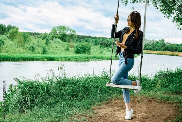 Fototapeta A happy young girl is standing on a swing in a beautiful tree by the river.