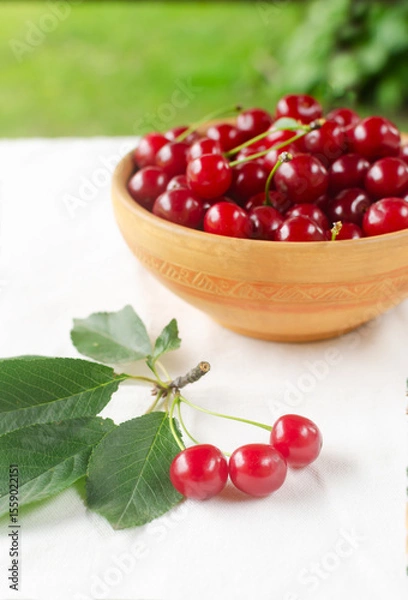 Fototapeta Ripe red cherries in a ceramic bowl with a branch on a white background. Concept of summer berry harvest. Vertical orientation. Selective focus.