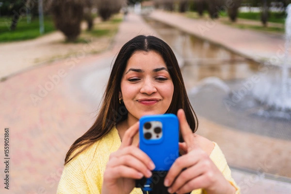 Fototapeta Young woman creating content for social media using a smartphone in a park