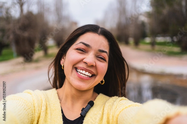 Fototapeta Young influencer smiling and creating content for social media, recording a video with a lavalier microphone in a park