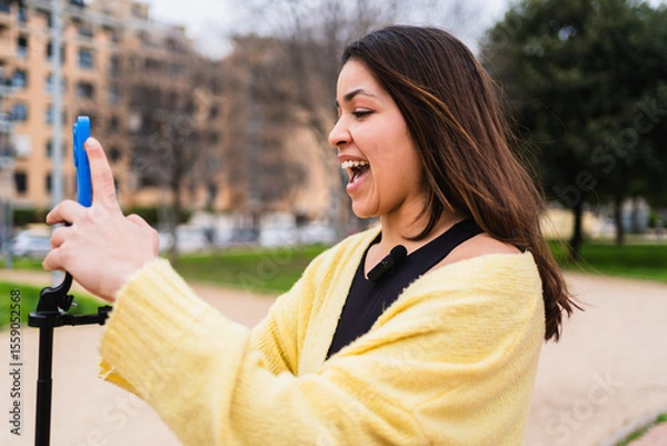 Fototapeta Young woman vlogger recording video for social media with her phone on a tripod in a park