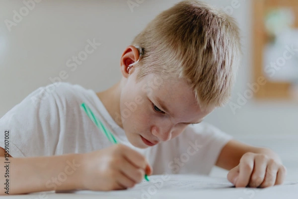 Fototapeta A boy with hearing aids focuses on homework, showcasing natural inclusive education. Authentic learning moment without staged intervention, perfect for disability awareness content