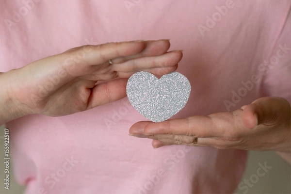 Obraz Close-up of female hands holding a shiny silver heart on a soft pink background, symbolizing love, kindness, and emotion