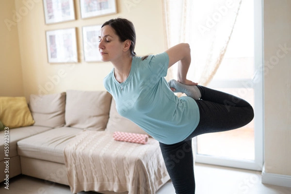 Obraz Woman in yoga clothes doing a standing quad stretch in a cozy living room. She is focused and balanced, practicing fitness at home