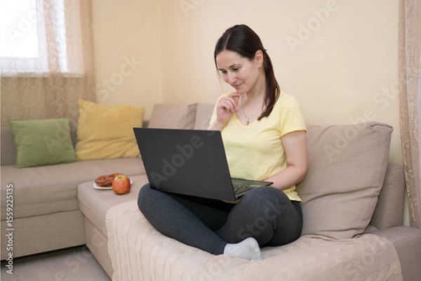 Obraz Woman sitting cross-legged on a sofa, focused on a laptop. An apple lies next to her. She is working or studying