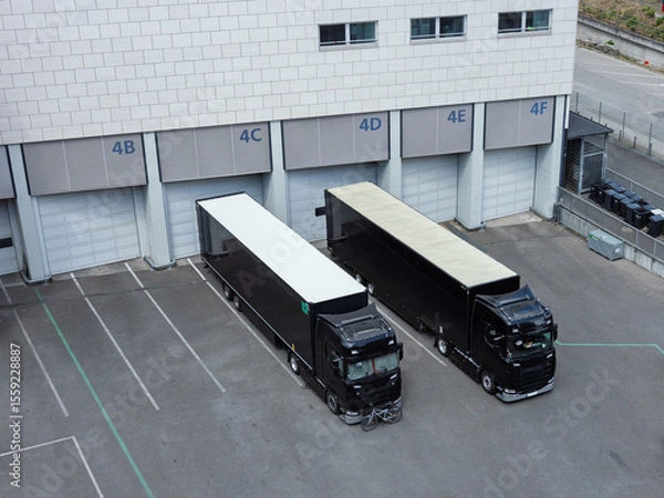 Fototapeta Two large black semi-trucks are parked at a loading dock with numbered bays in an industrial area, viewed from above