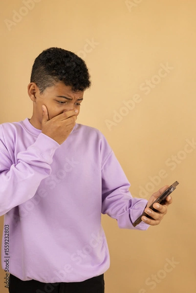 Fototapeta Vertical image of a young brown skinned Latino man wearing a lilac sweatshirt with a horrified expression with his hand over his mouth when seeing something on his cell phone on a brown background