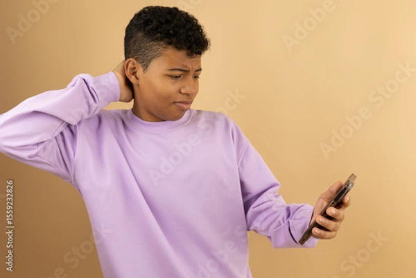 Fototapeta Young brown skinned Latino man wearing a lilac sweatshirt with a disgusted expression when seeing something on his cell phone on a light brown background with copyspace