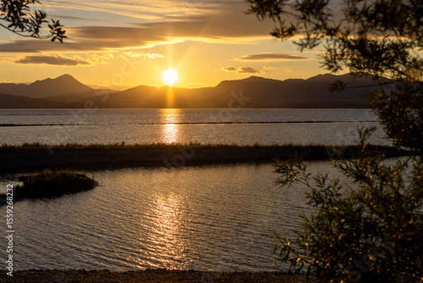 Fototapeta Beautiful sunset over the mountains above the lake. Reflection of sun rays in the water in a sea bay with trees