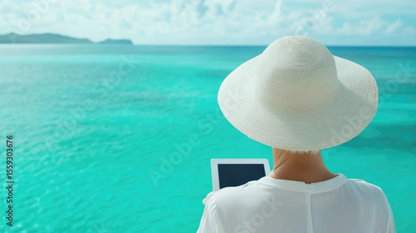 Fototapeta A person in a wide-brimmed hat looks at a tablet while facing a vibrant turquoise sea. Relaxation, technology, and ocean view