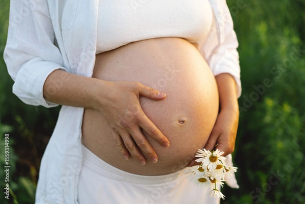 Fototapeta A pregnant woman stands in a green field. She gazes lovingly at her baby bump as the sun sets behind her, creating a serene atmosphere.