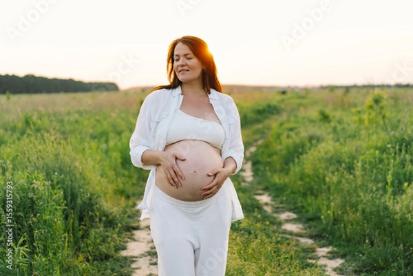 Fototapeta A pregnant woman stands in a green field. She gazes lovingly at her baby bump as the sun sets behind her, creating a serene atmosphere.