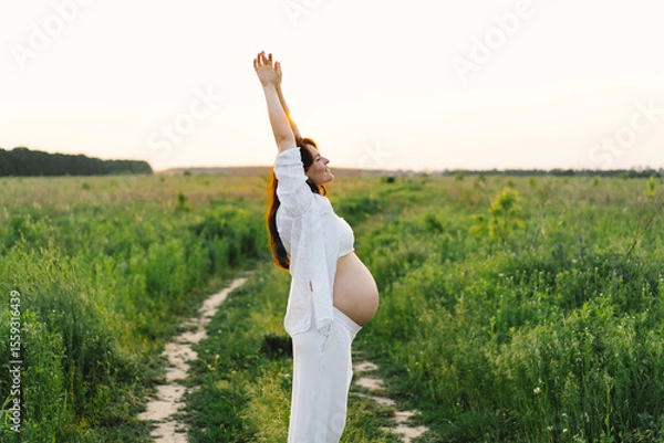 Fototapeta A pregnant woman stands in a green field. She gazes lovingly at her baby bump as the sun sets behind her, creating a serene atmosphere.