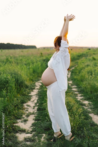 Fototapeta A pregnant woman stands in a green field. She gazes lovingly at her baby bump as the sun sets behind her, creating a serene atmosphere.