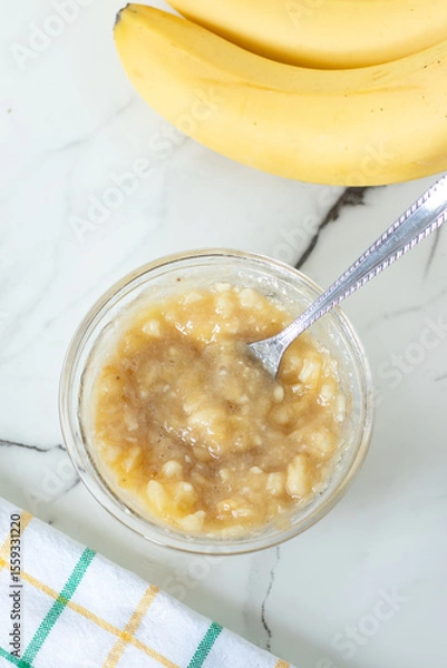 Obraz Mashed banana in a bowl with fork on white kitchen table. Top view.