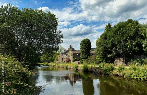 Fototapeta The Leeds and Liverpool Canal flows past an old stone house with leafy trees, mirrored in calm water beneath a dappled sky in Shipley, Yorkshire. UK