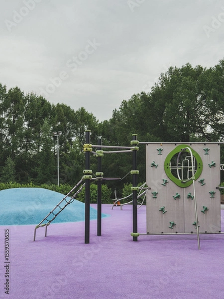 Fototapeta Colorful modern playground with climbing walls and structures on purple rubber surface and blue hill, surrounded by trees, concept of childhood activity and safe outdoor play