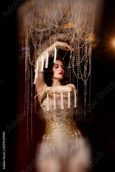 Fototapeta Elegant young circus actress in golden corset and gloves, standing under chandelier, front view. Retro theatrical look for circus, burlesque, performance, vintage stage shows.