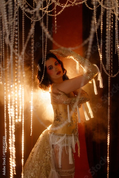 Fototapeta Young Caucasian circus performer or burlesque dancer in golden vintage corset, front view, posing under chandelier on dark background. Concepts: circus, cabaret, retro, show, performance.