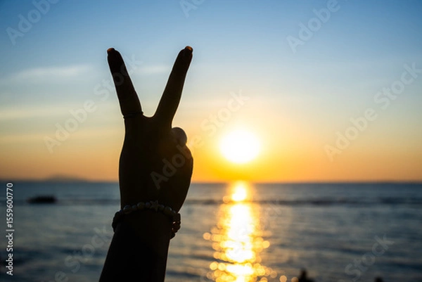 Obraz silhouetted hand making a peace sign against a glowing sunset over the ocean, with golden light