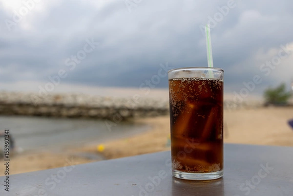 Obraz glass of iced cola sits on a table by the sea, with a cloudy sky backdrop. The drink's bubbles and condensation are visible