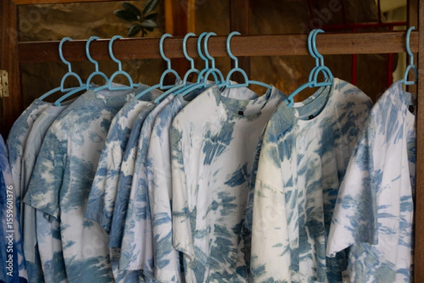 Obraz close-up shot of tie-dye t-shirts hanging on light blue plastic hangers. The shirts feature a soft, pastel tie-dye pattern.