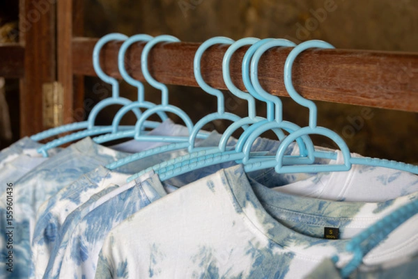 Obraz close-up shot of tie-dye t-shirts hanging on light blue plastic hangers. The shirts feature a soft, pastel tie-dye pattern.
