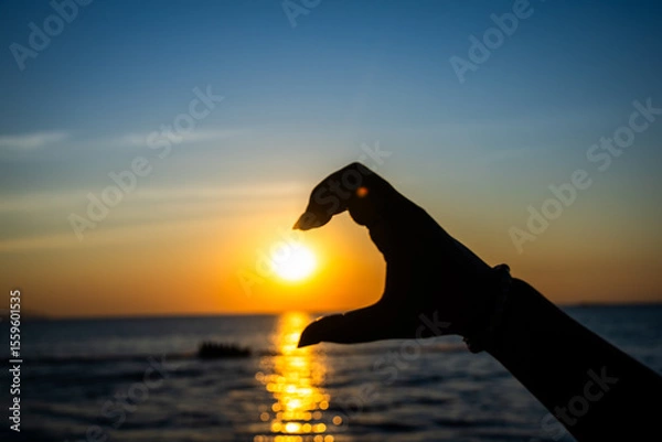 Obraz silhouetted hand forming a half-heart gesture in front of a glowing sunset over the ocean