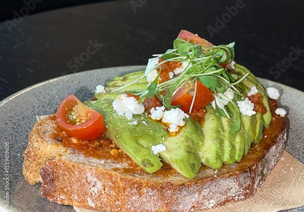 Fototapeta Avocado toast bread, slices garnished with tomatoes and feta cheese, microgreens and sauce, in ceramic plate on black wood table, high angle view, copy space