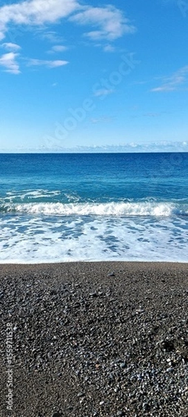 Fototapeta Pebble beach with gentle waves washing ashore under a bright blue sky with scattered clouds