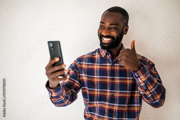 Fototapeta Ecstatic black man in cozy shirt standing in front of wall and having video call on smartphone.