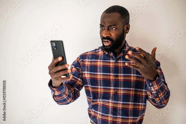 Fototapeta Angry black man in cozy shirt standing in front of wall and having video call on smartphone. He is arguing with someone.