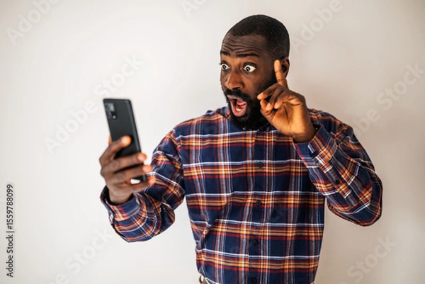 Fototapeta Surprised and shocked black man in cozy shirt standing in front of wall and having video call on smartphone.