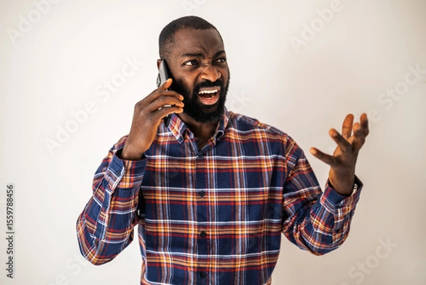 Fototapeta Angry black man in cozy shirt standing in front of wall and talking on mobile phone.