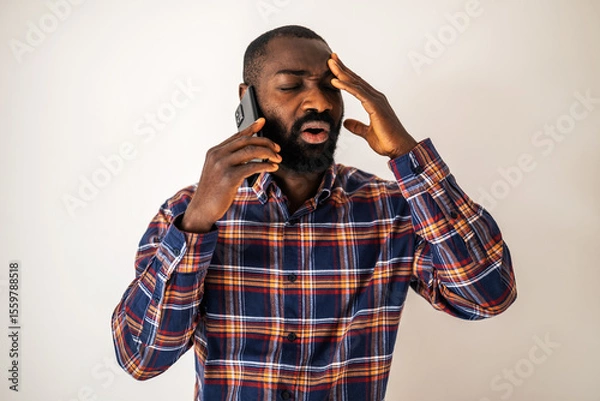 Fototapeta Anxious and tired black man in cozy shirt standing in front of wall and talking on mobile phone.