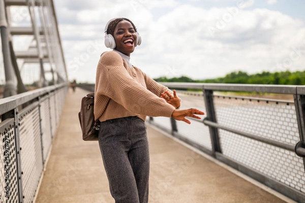 Fototapeta Happy young black woman is walking on bridge foot path, listening music and smiling. She is enjoying sunny day outdoor.