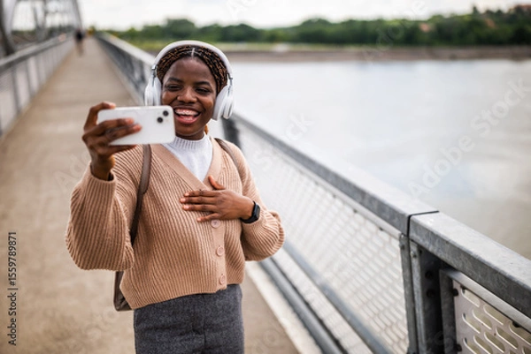 Fototapeta Happy young black woman is looking at her phone and smiling. She is enjoying sunny day outdoor and listening music while using smartphone.